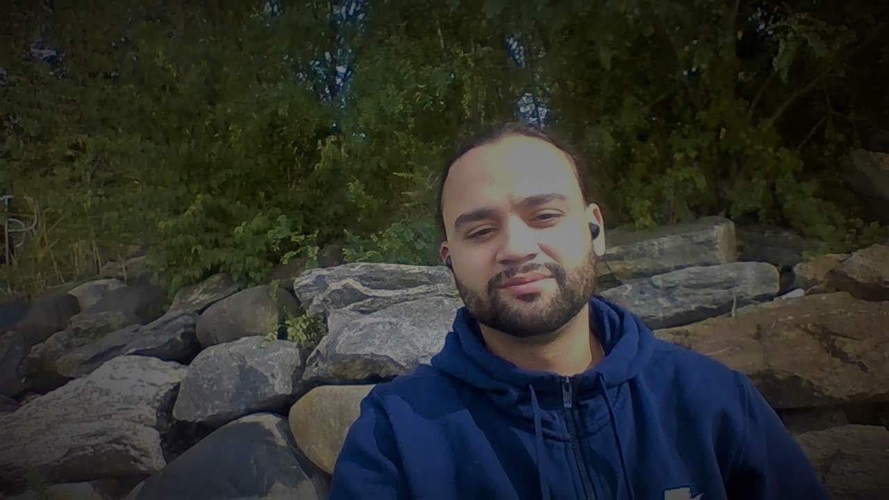 Gio outdoors, sitting beside rocks and trees in a blue hoodie